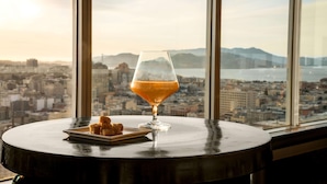 Una mesa con un vaso de líquido ámbar, un plato de pequeños pasteles y vistas panorámicas de la ciudad a través de amplias ventanas.