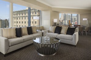 A modern hotel room with two sofas facing a large window, a glass coffee table, and a city view.