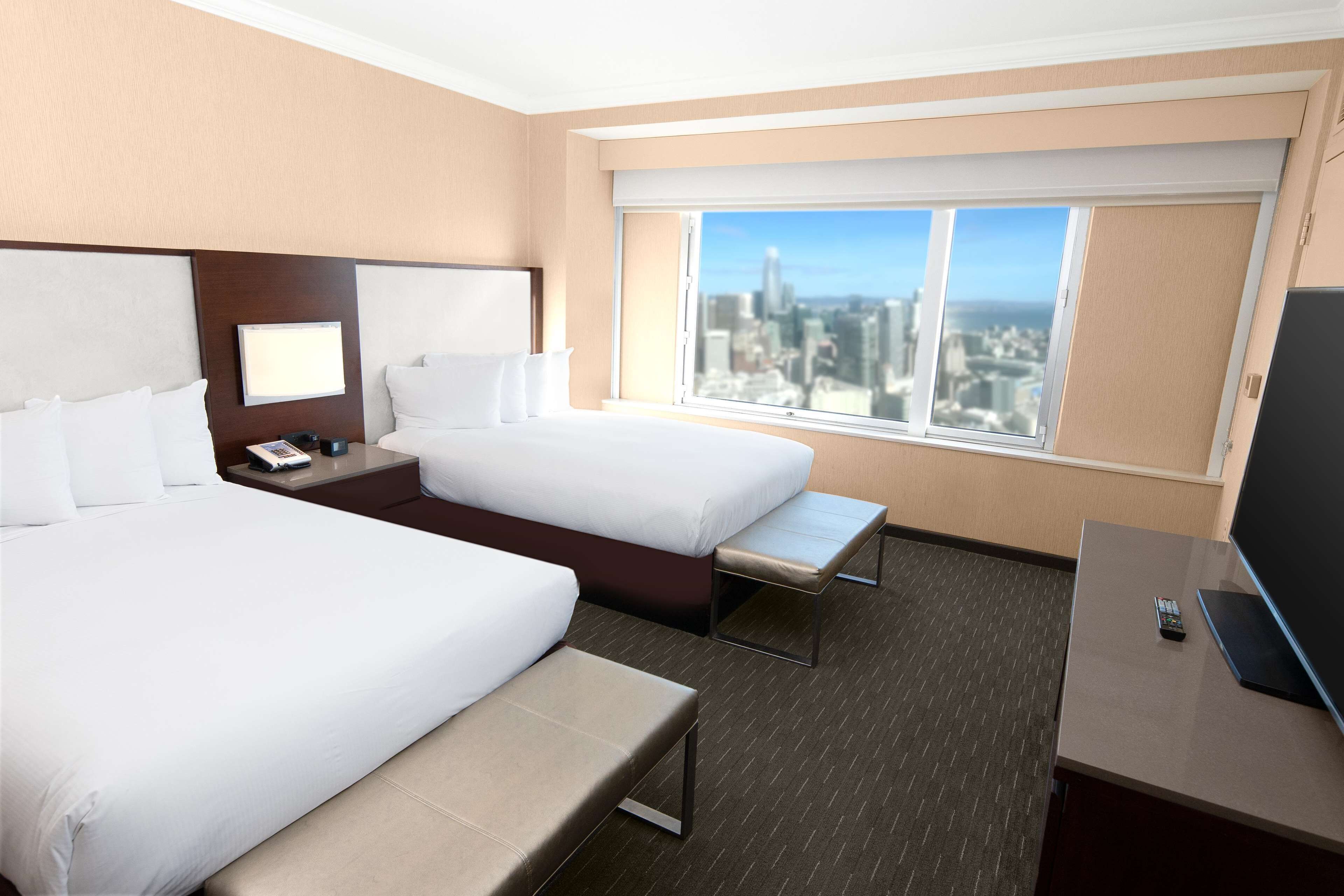 Habitación de hotel con dos camas, un escritorio y un amplio ventanal con vista a la ciudad.