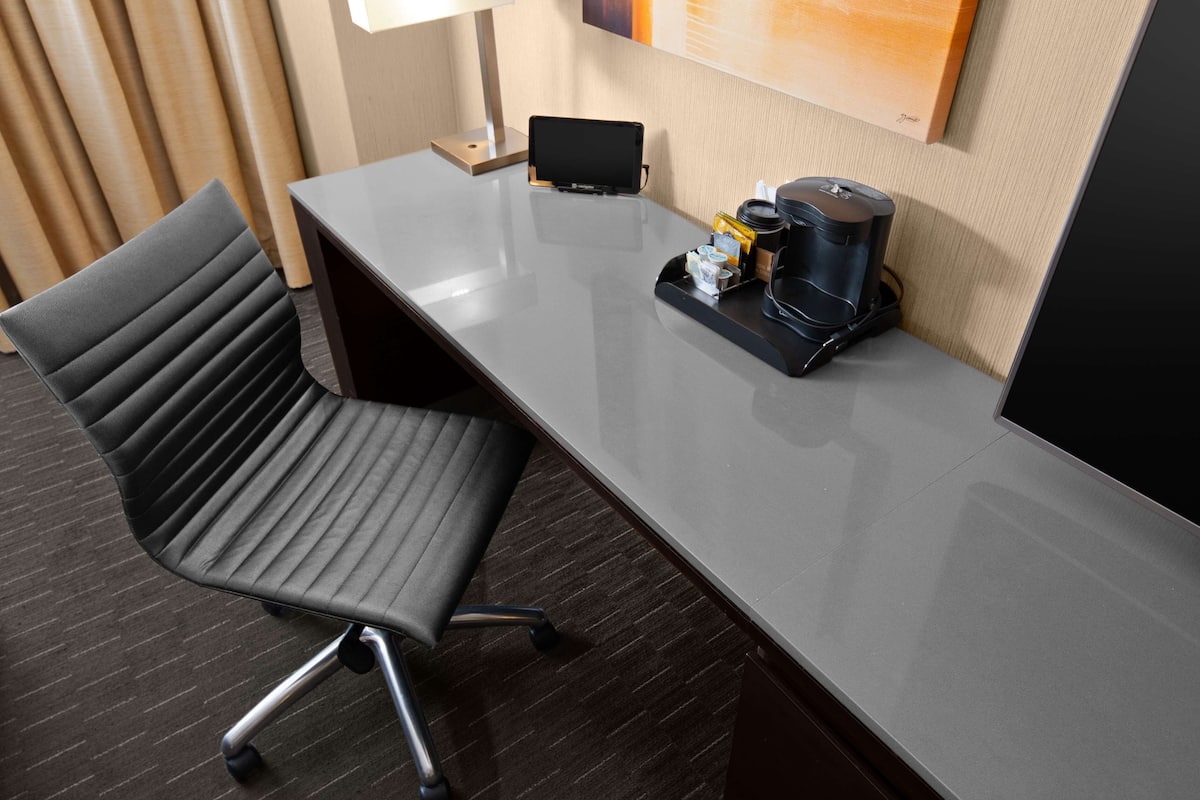 Una habitación de hotel con un escritorio moderno, una silla de oficina negra, una cafetera y una computadora portátil sobre el escritorio.