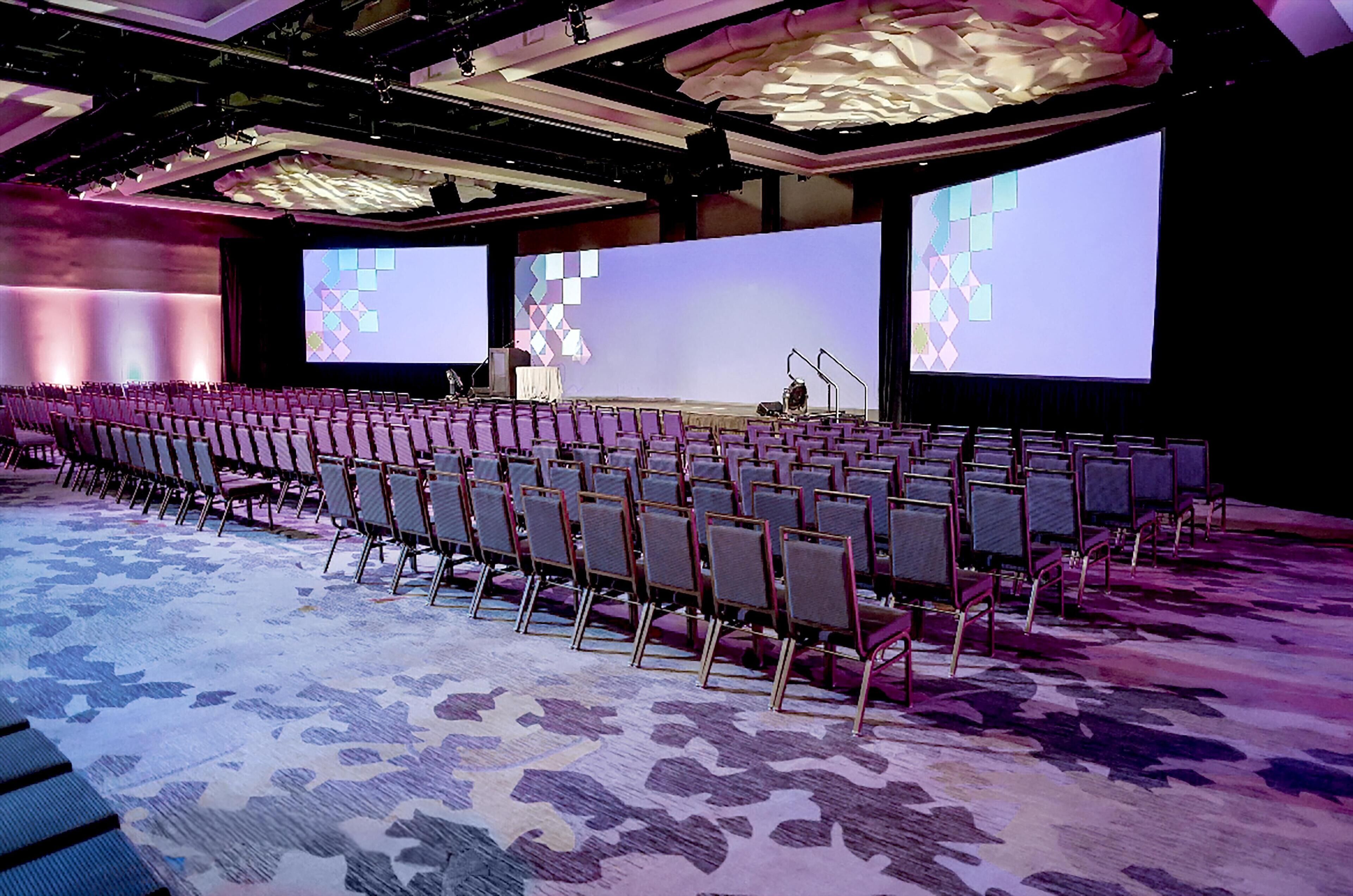 A conference room with rows of chairs, a stage with a large screen, and a patterned carpet.
