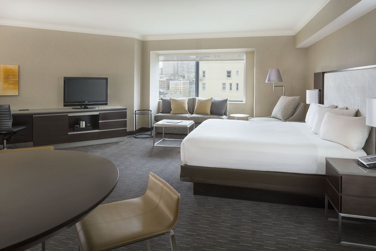 Habitación de hotel con una cama grande, un escritorio con televisor de pantalla plana, un sofá, una mesa de comedor y vistas a la ciudad por la ventana.