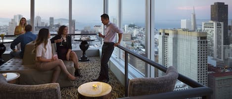 A high-rise hotel room with a city view, large windows, and a balcony with seating.