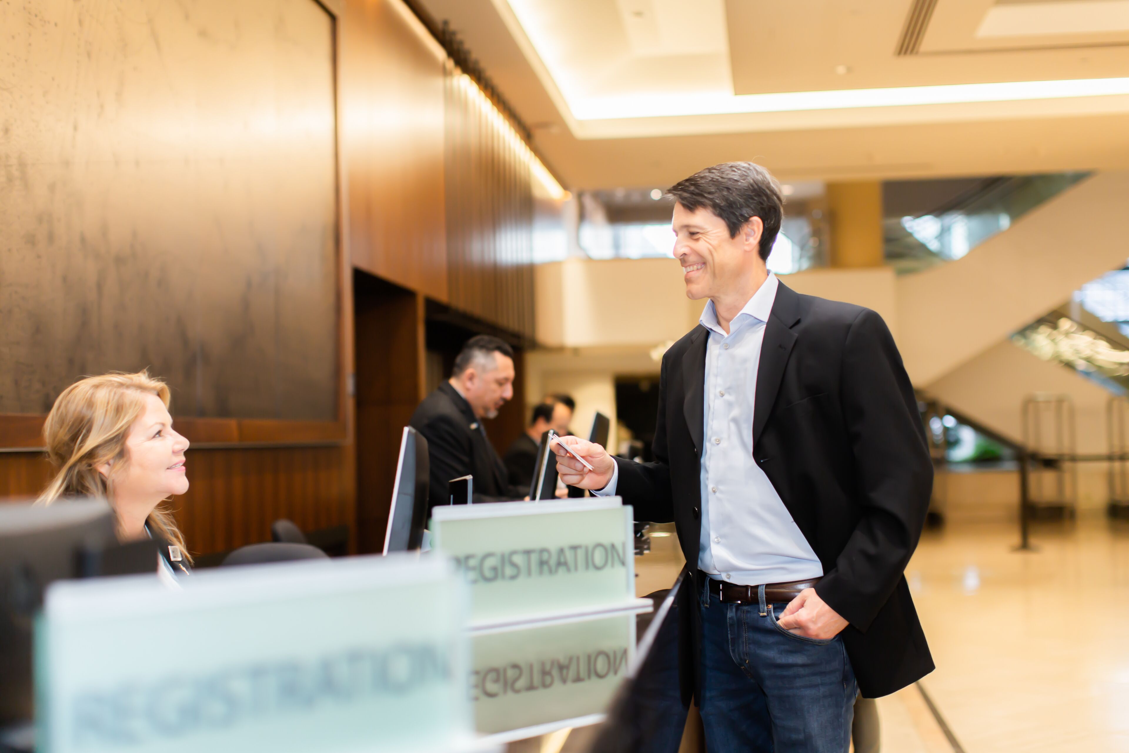 A man in a black blazer and jeans standing at a registration desk in a modern lobby with wooden paneling and a glass ceiling.