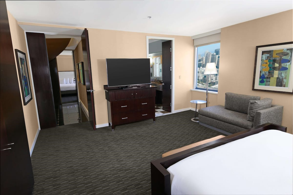 Habitación de hotel con una cama, un televisor sobre una cómoda, un sofá gris, una ventana con vista a la ciudad y una puerta que da a otra habitación.