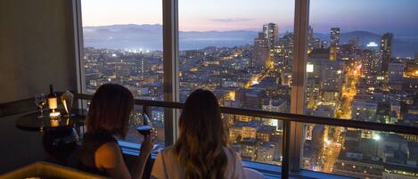 Vista de um quarto em andar alto com vista para a cidade ao anoitecer.