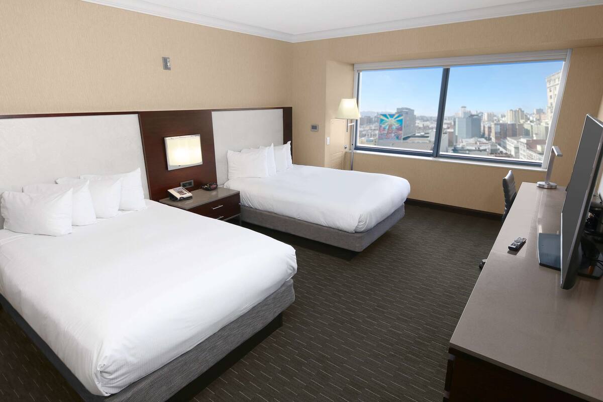 Habitación de hotel con dos camas, un escritorio, un televisor y una ventana con vista a la ciudad.