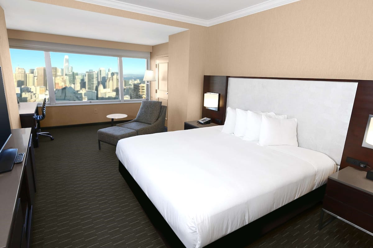 Habitación de hotel con una cama grande, un escritorio con una computadora portátil, una silla, una mesita y vistas a un horizonte urbano a través de un ventanal amplio.