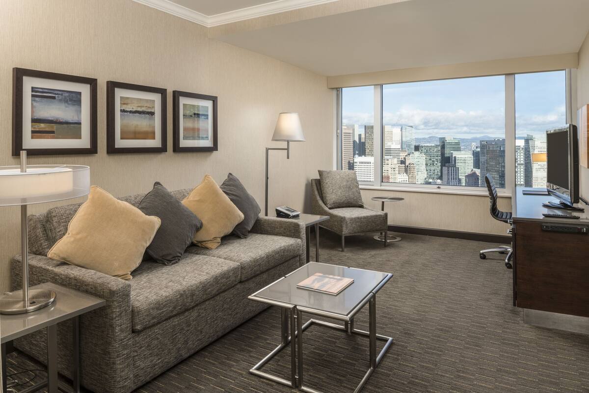 Una habitación de hotel moderna con un sofá gris, una mesa de centro de vidrio, un escritorio con una computadora y un amplio ventanal con vistas a la ciudad.