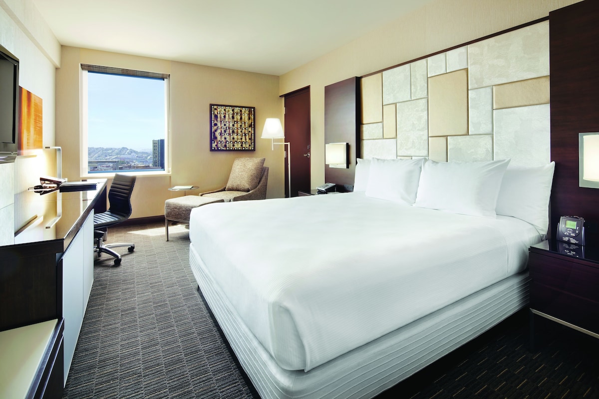 A hotel room with a large bed, a desk, a chair, a TV, a window with a city view, and a decorative wall panel.