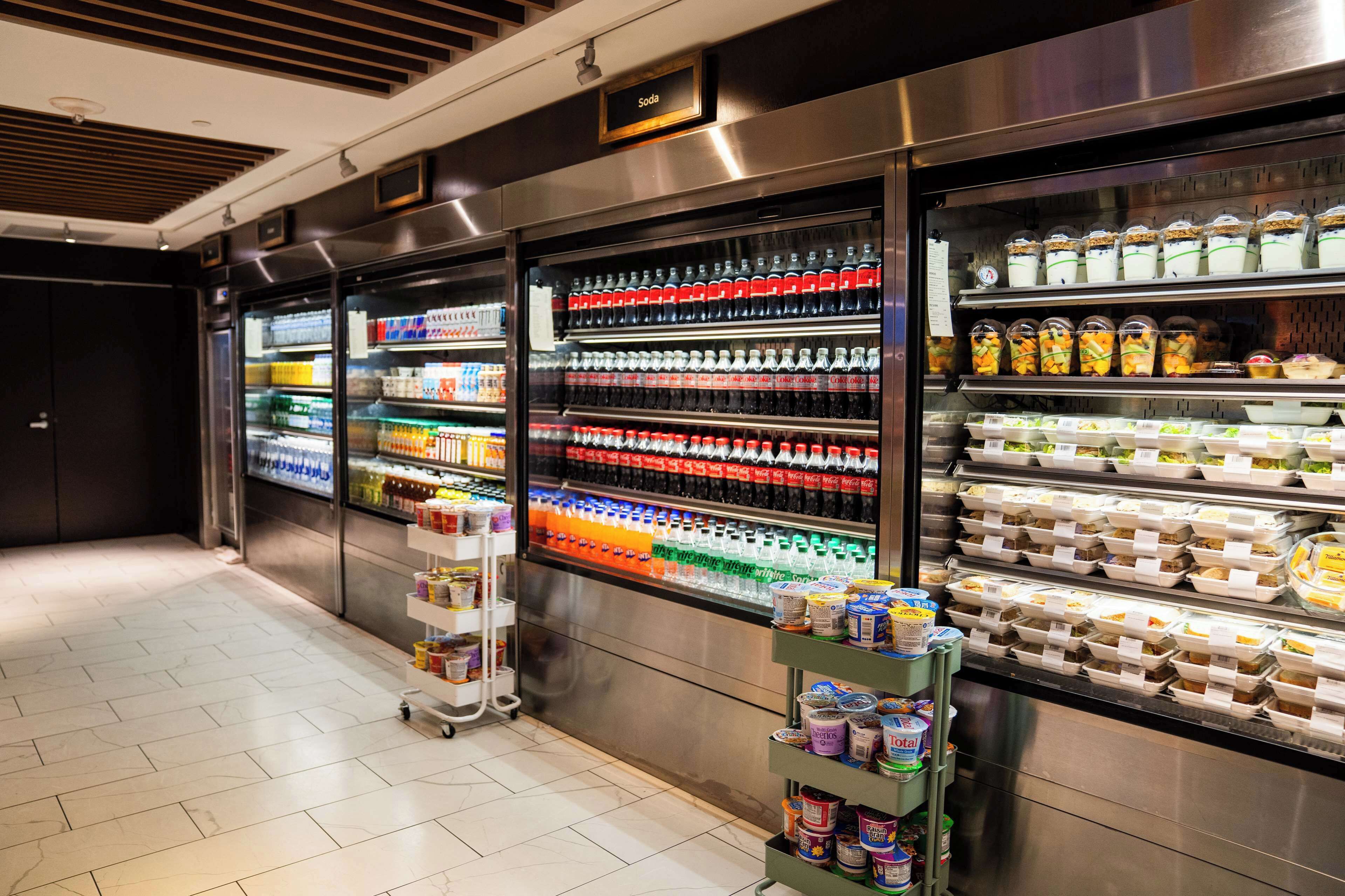 Een goed verlichte, moderne ruimte voor catering, met gekoelde drankdispensers, diverse verpakte etenswaren en een zelfbedieningsdrankenwagen.