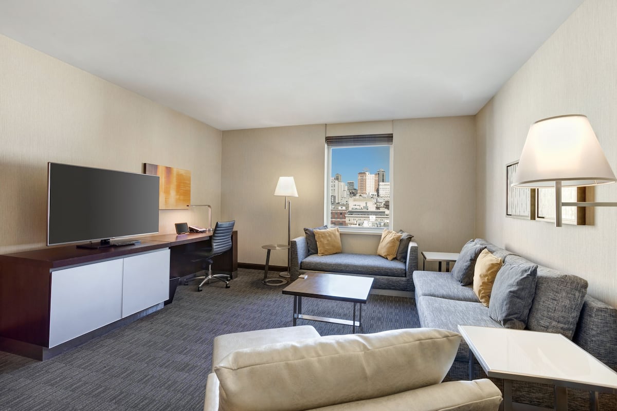 Habitación de hotel con televisión de pantalla plana, escritorio con silla, sofá, mesa de centro y lámpara.