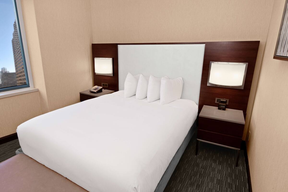 Una habitación de hotel con una cama grande, dos mesitas de noche con lámparas, una mesita auxiliar y una ventana con vistas a la ciudad.