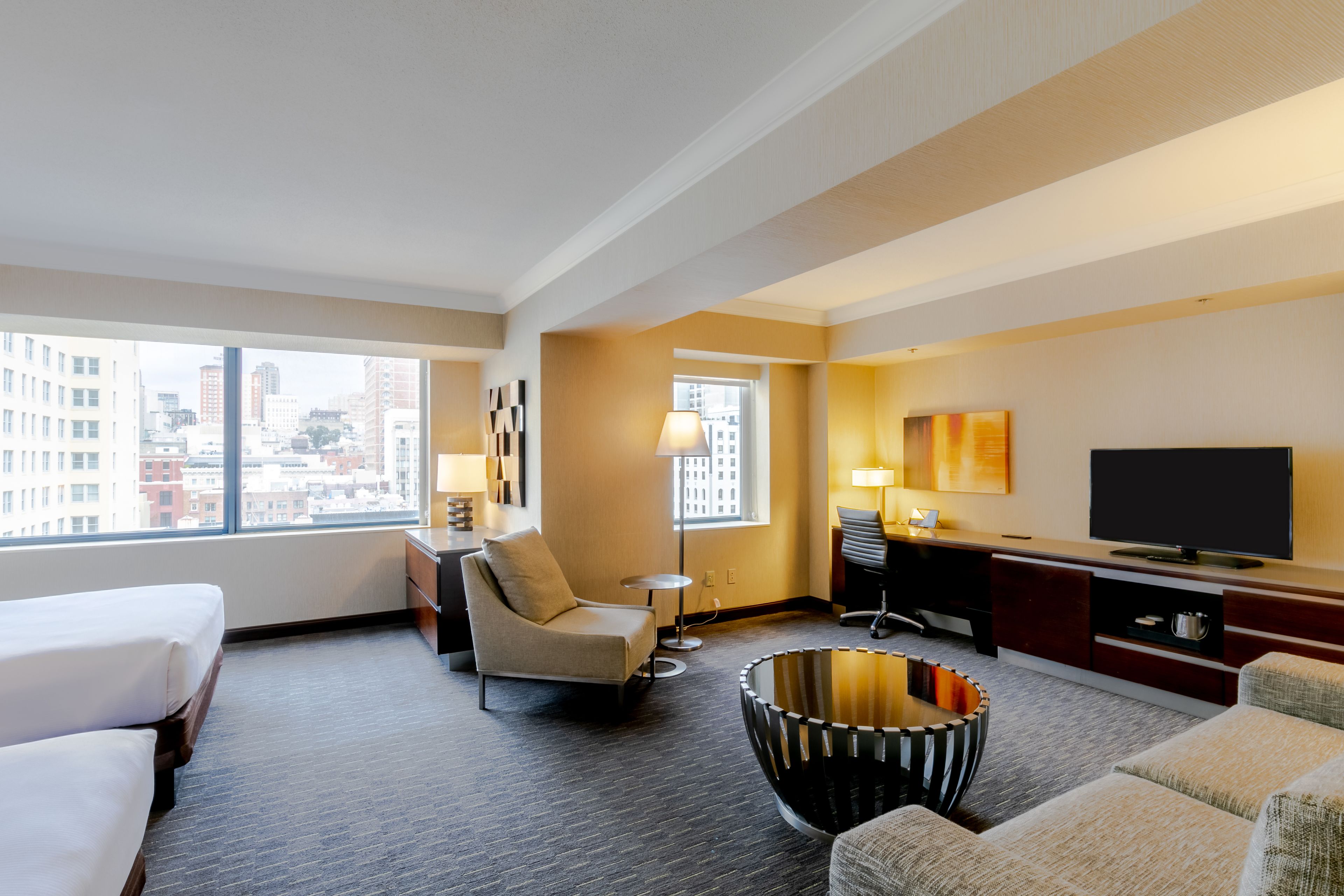 A hotel room with a bed, a sofa, a chair, a TV, a lamp, and a view of the city.