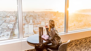 Una mujer sentada junto a una ventana con vista a la ciudad, sosteniendo una copa de vino y un bolso rosa.