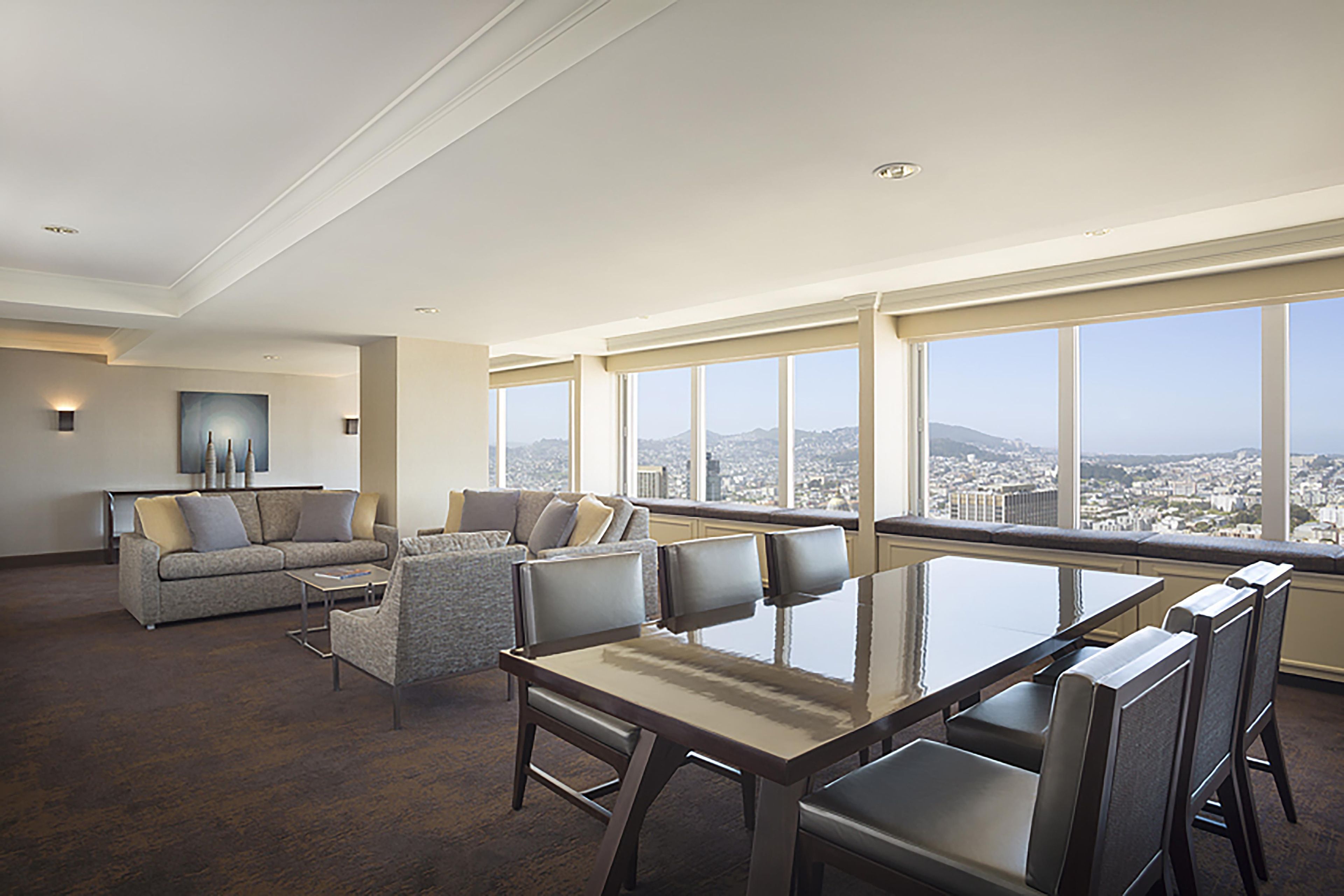A spacious living area with a high ceiling, a dining table with chairs, a sofa set, and a large window offering a city view.