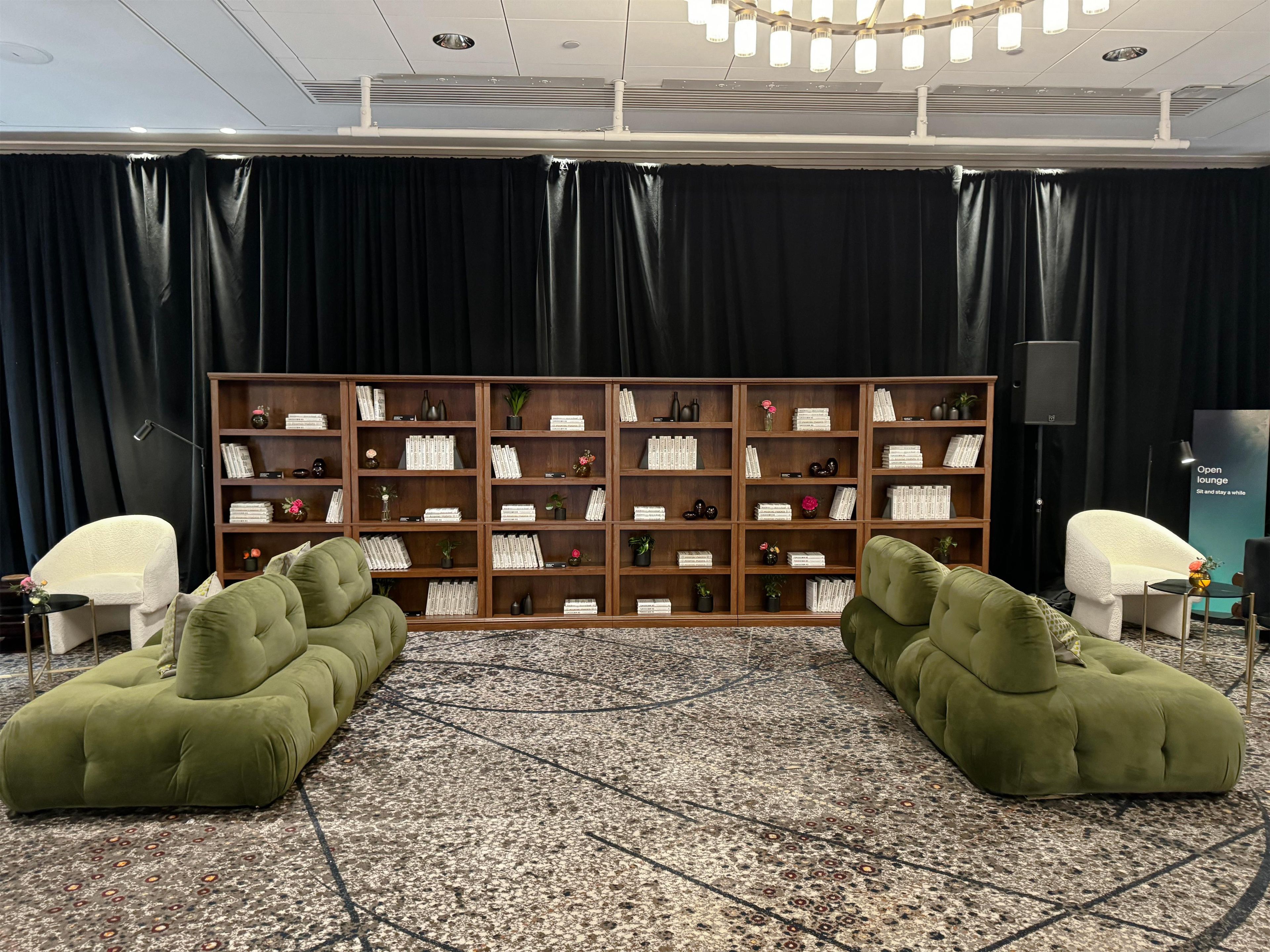 A room with a large bookshelf, two green sofas, a patterned carpet, and a black curtain backdrop.
