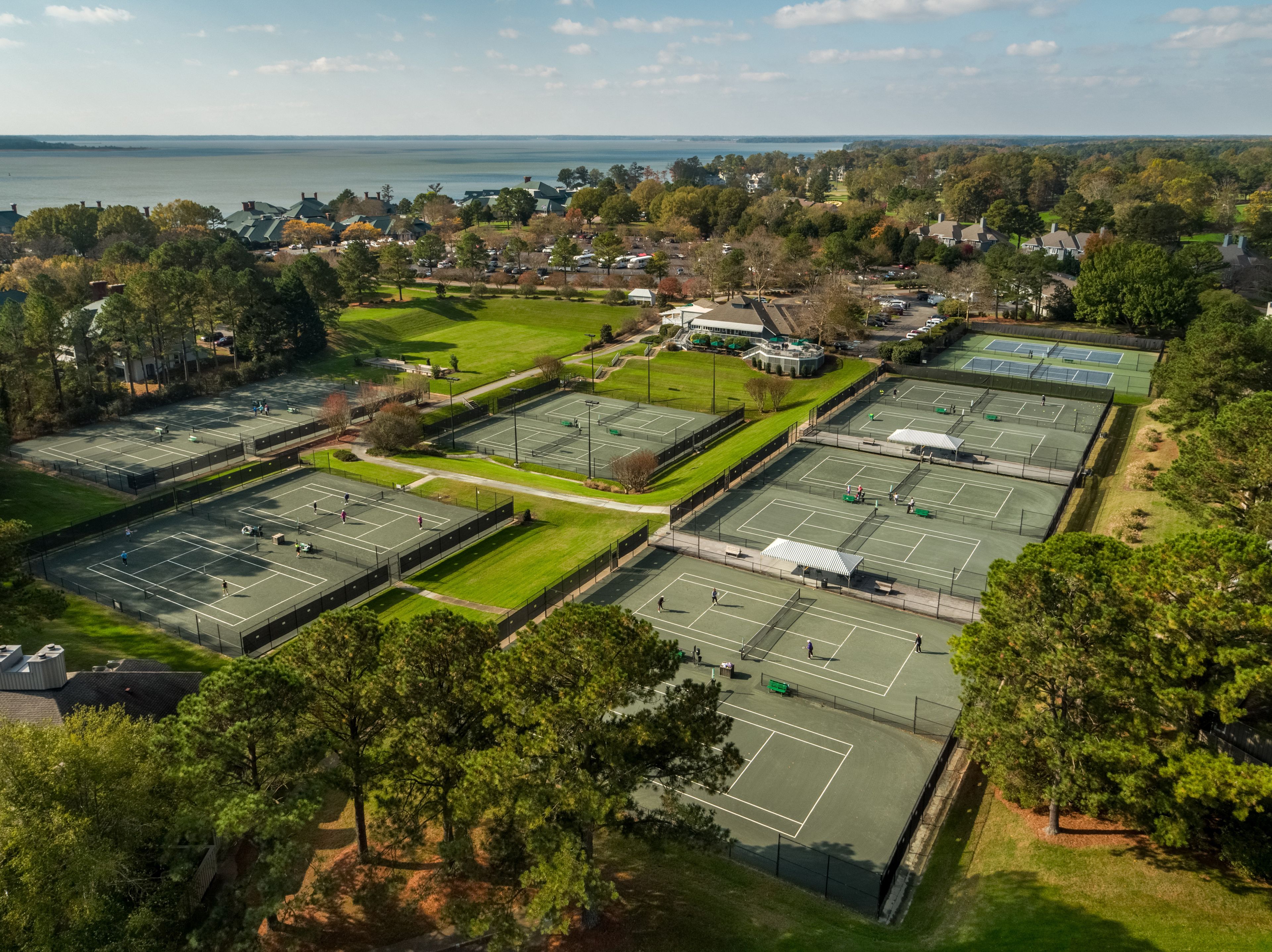 An aerial view of a tennis facility with multiple courts, surrounded by trees and residential buildings.
