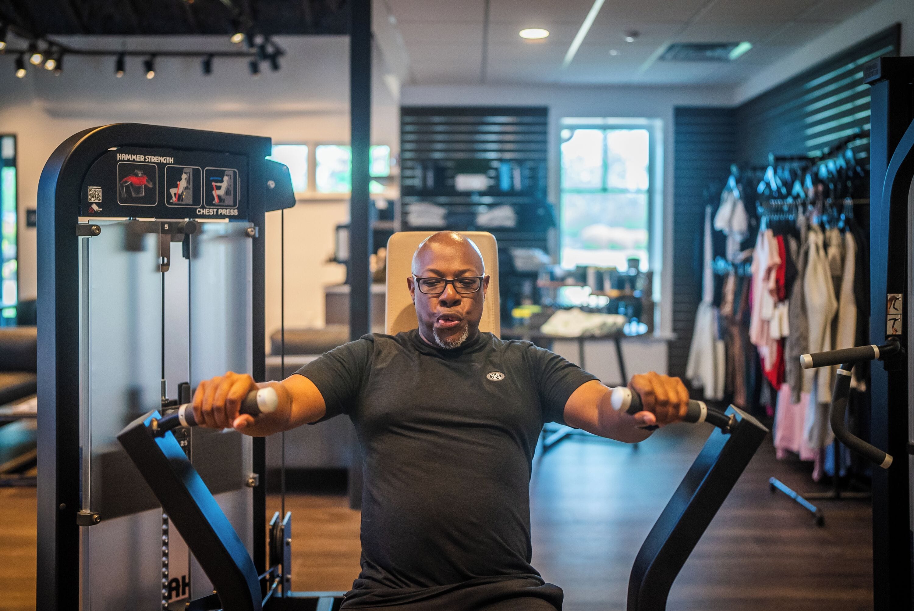 A man using a chest press machine in a gym with a clothing rack in the background.