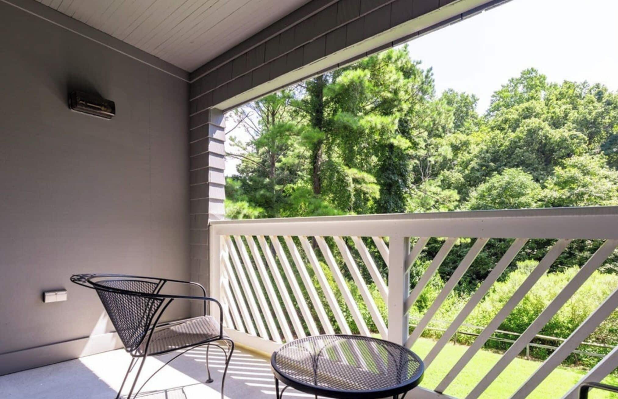 Een balkon met een witte railing, twee metalen stoelen en uitzicht op een groen landschap met bomen.