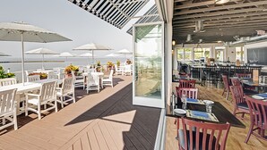 An outdoor dining area with tables and chairs, a view of the sea, and a glass door leading to an interior space.