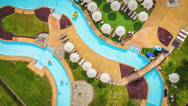 An aerial view of a resort with a large swimming pool, a water slide, and multiple lounge areas with umbrellas.