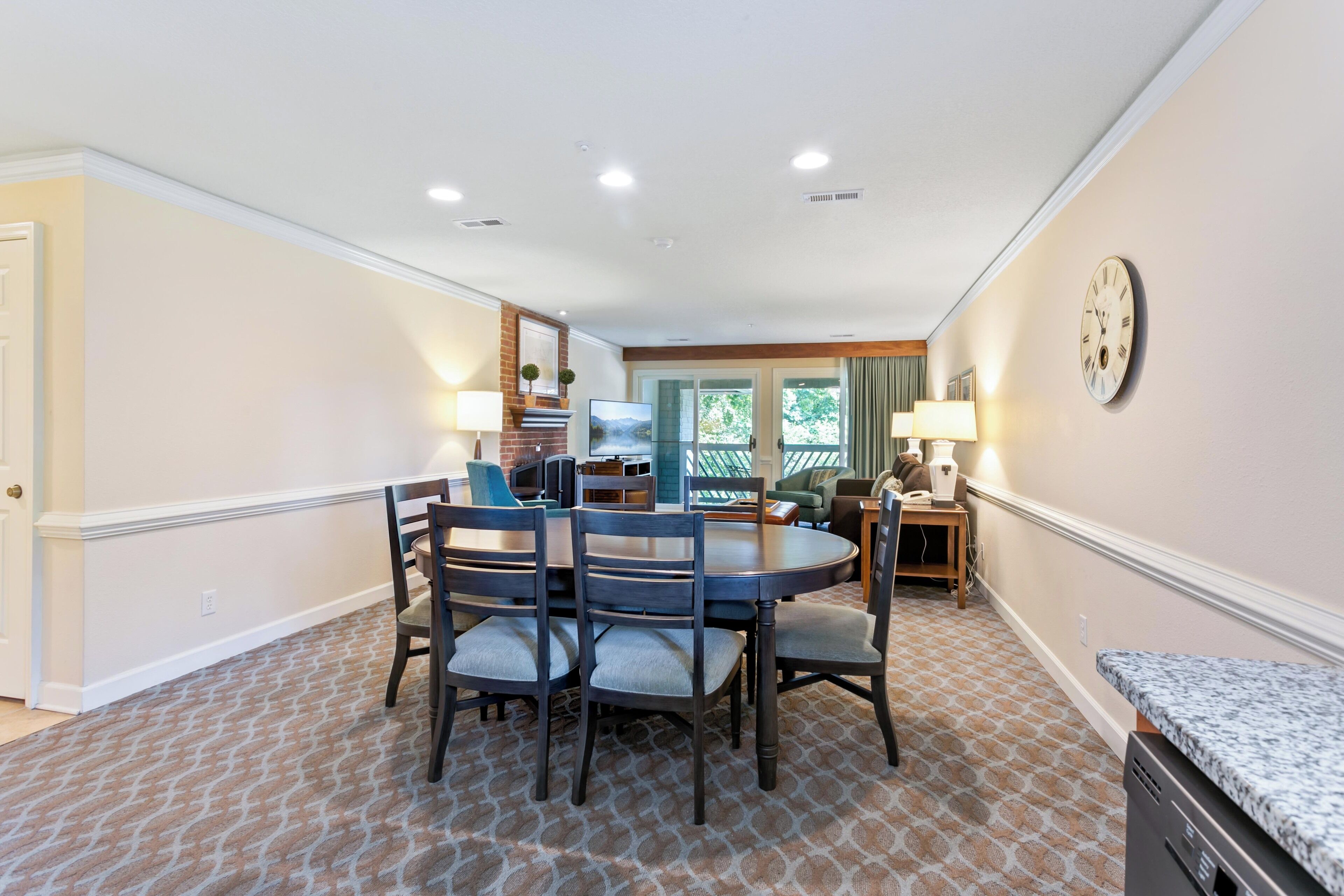 A dining area with a round table and chairs, a clock on the wall, and a view of the outdoors through a door.