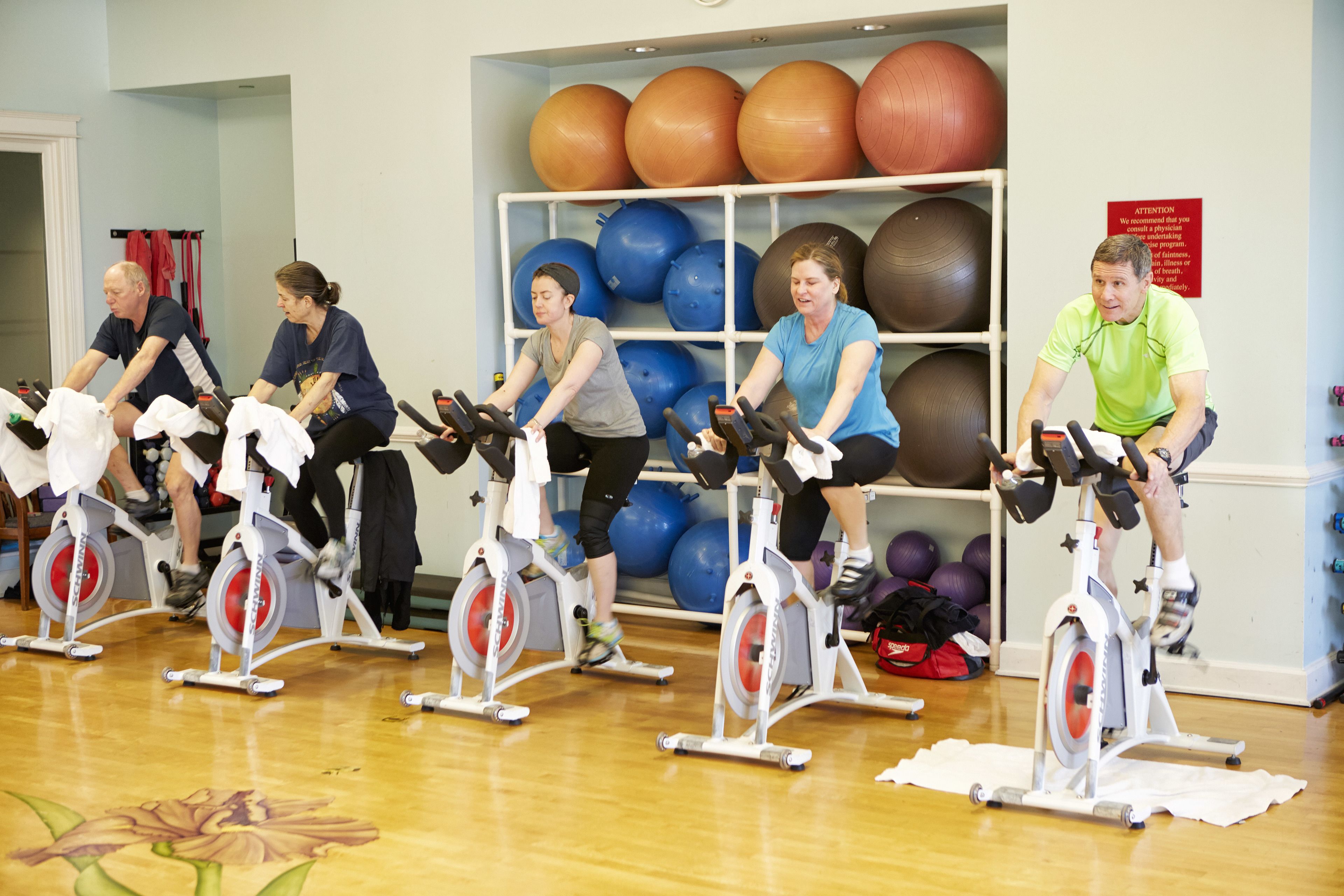 A group of people exercising on stationary bikes in a room with exercise balls on shelves.