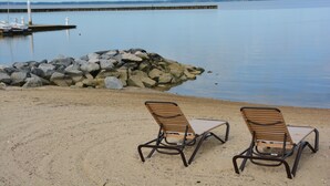 Two lounge chairs on a sandy beach with a rocky breakwater and calm water in the background.