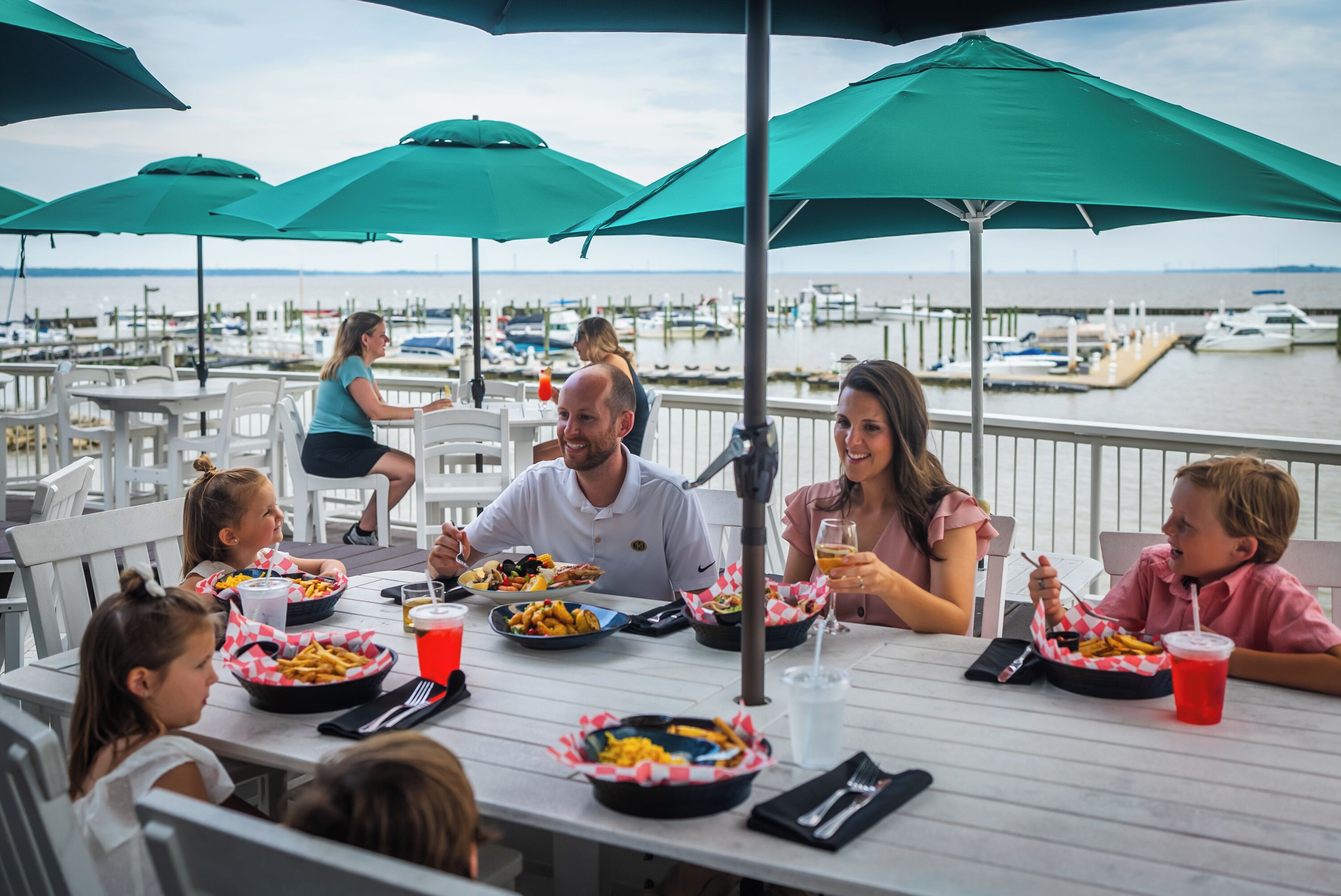 A dining area with tables and chairs, a view of the marina with boats, and umbrellas providing shade.
