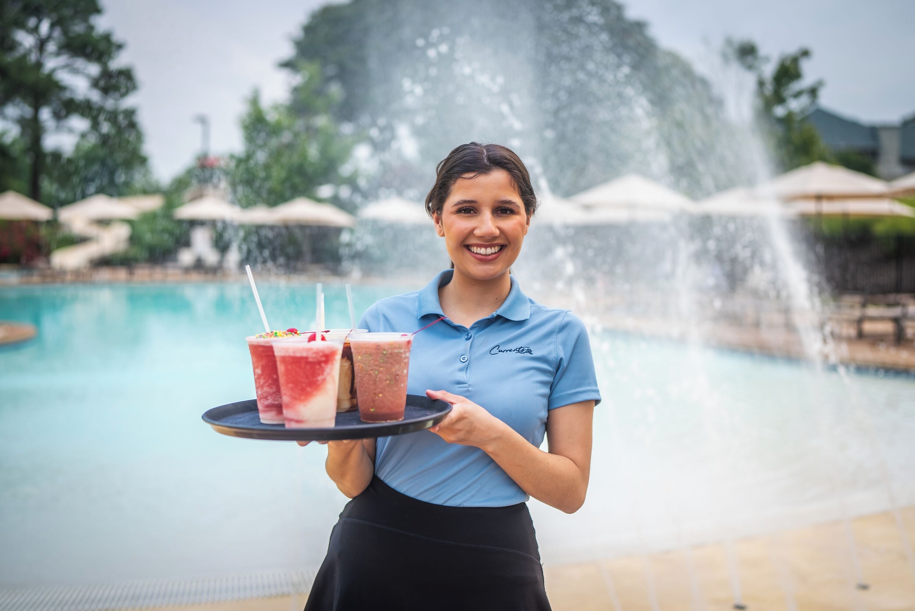 A person holding a tray with two drinks near a pool with a fountain.