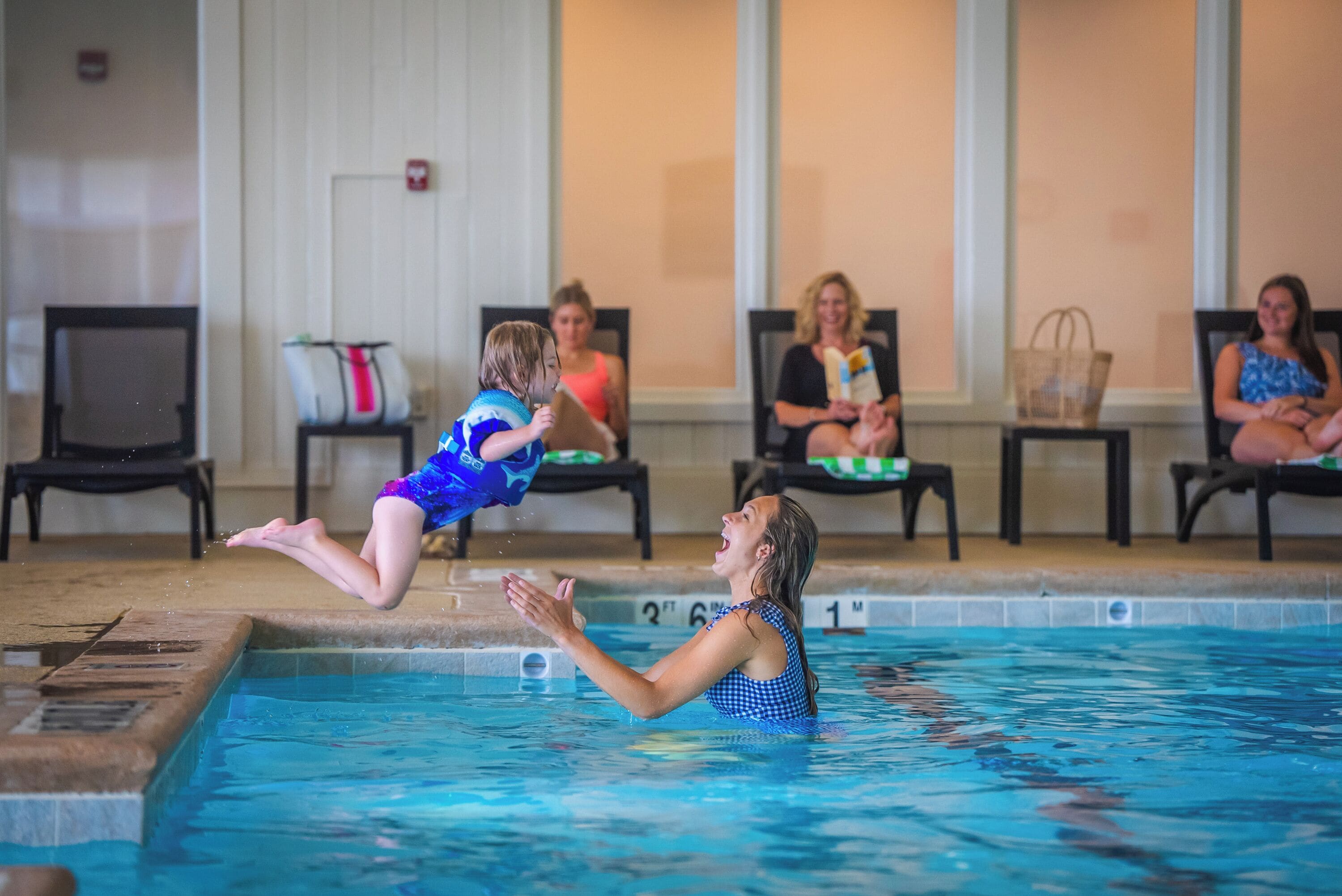 A swimming pool with a diving board, chairs, and a tiled floor.