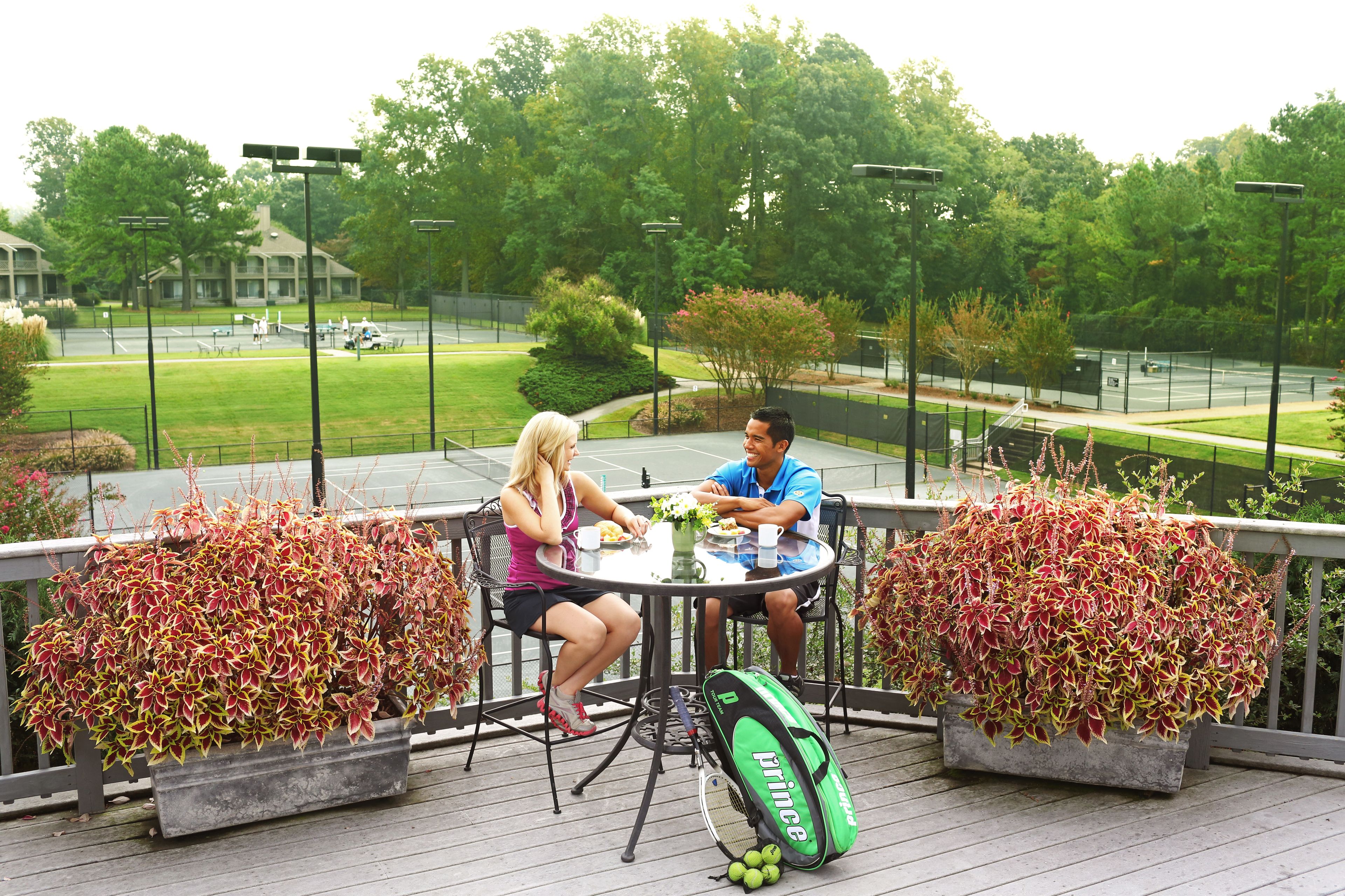 Two people dining at an outdoor table with potted plants on either side, overlooking a tennis court and greenery.