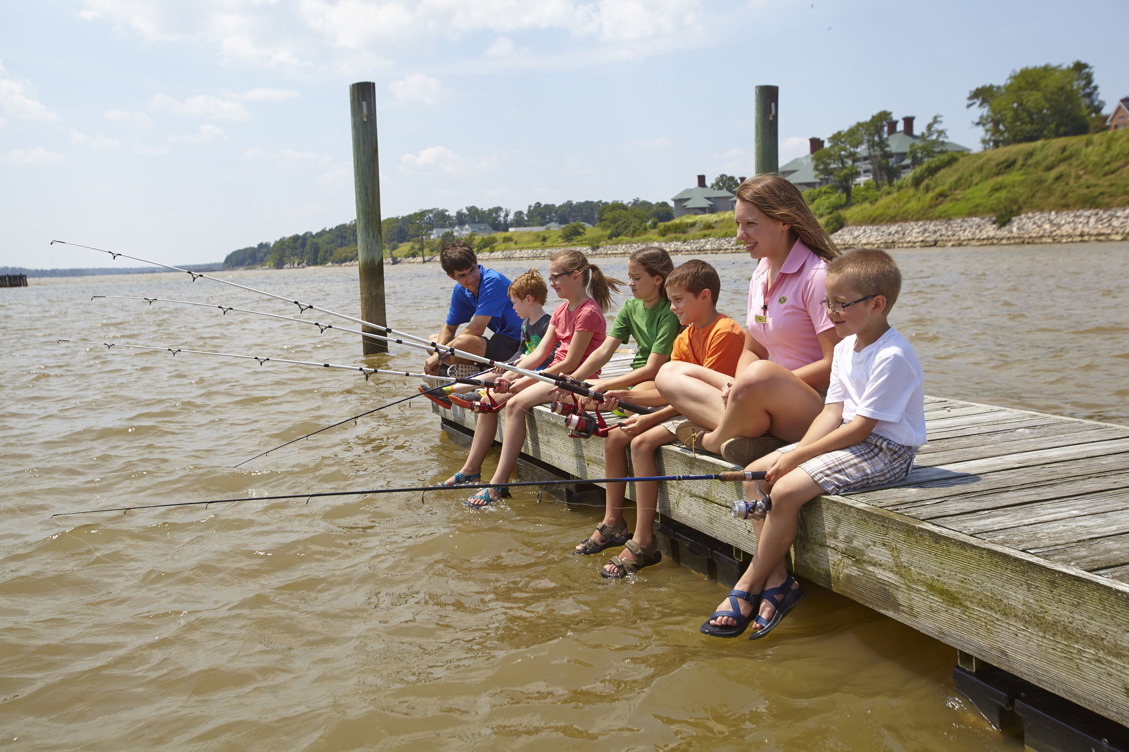A group of people sitting on a wooden dock by a river, holding fishing rods.