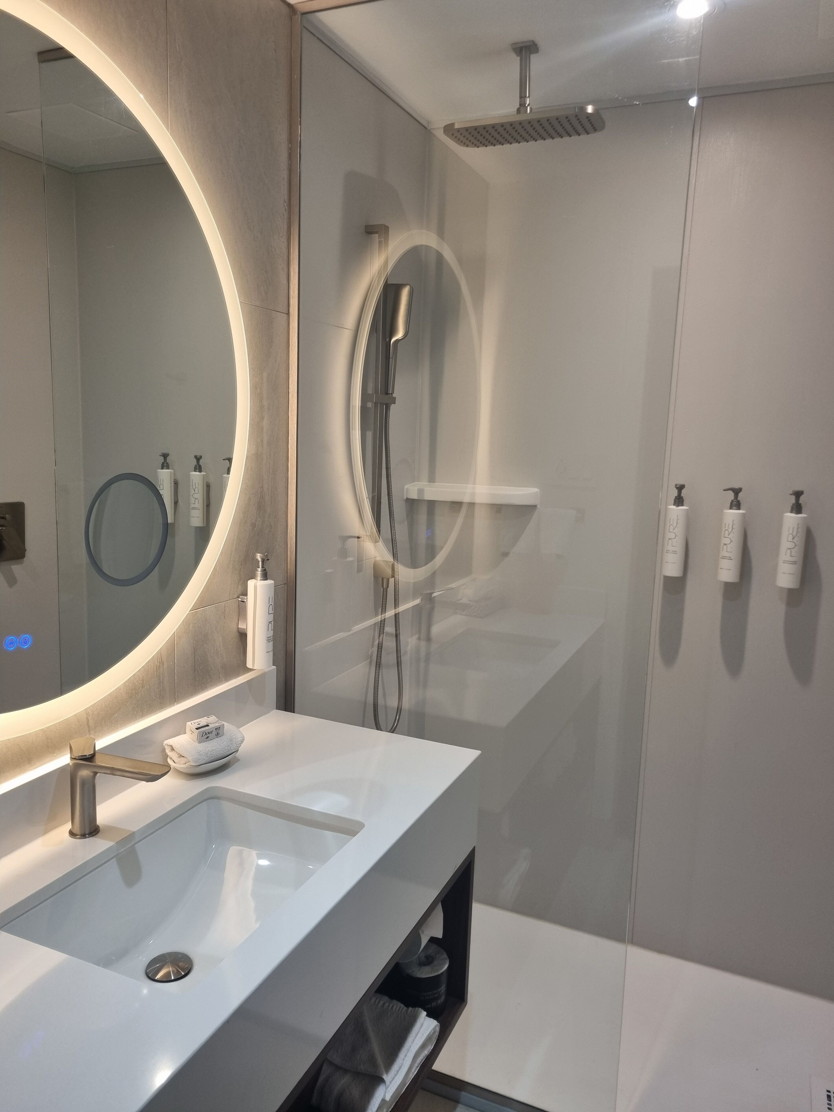 A modern bathroom with a large oval mirror, a white sink, and a glass-enclosed shower area.