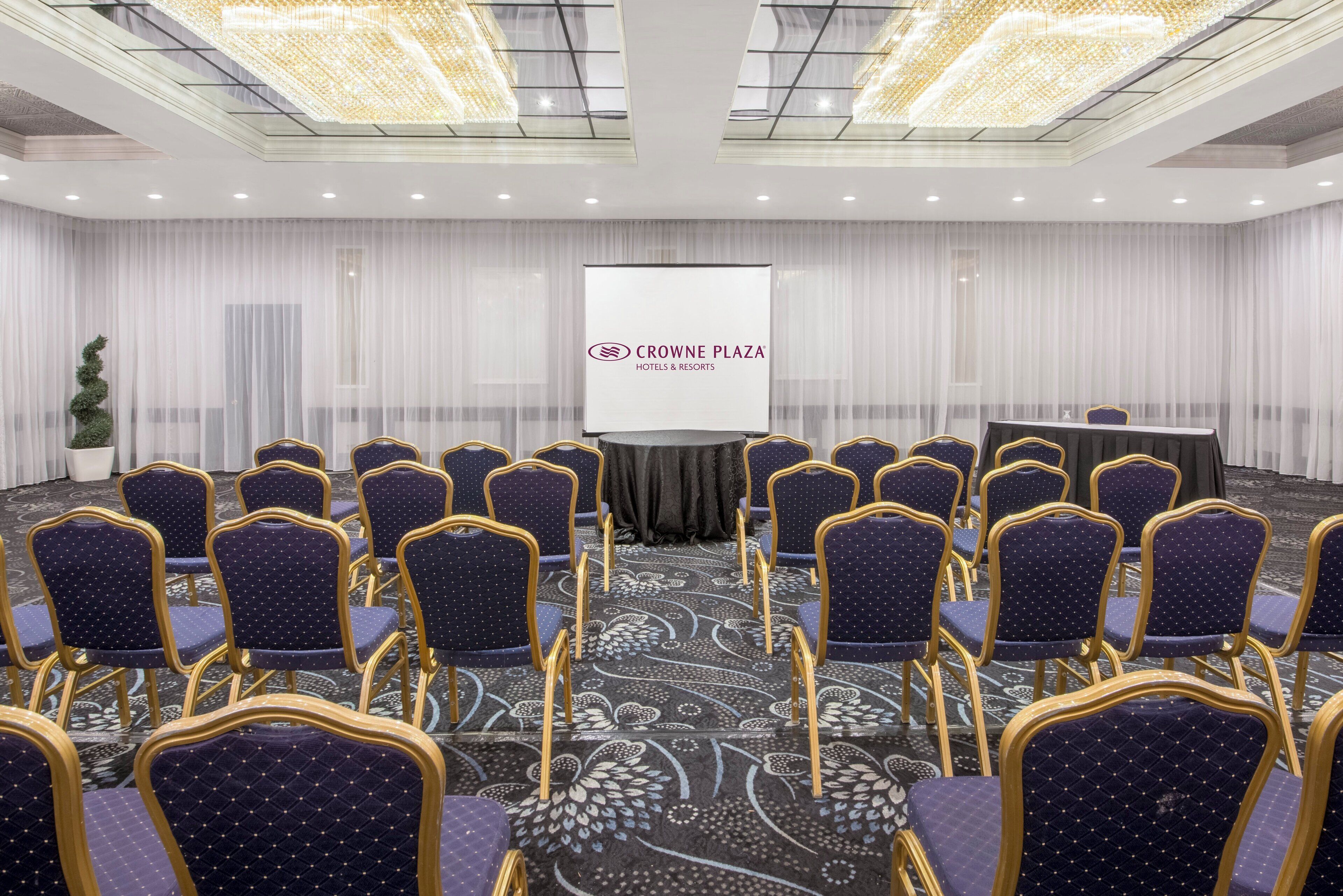 Un salón de conferencias con filas de sillas, un atril y una pantalla que muestra el logotipo de Crowne Plaza Hotels & Resorts.