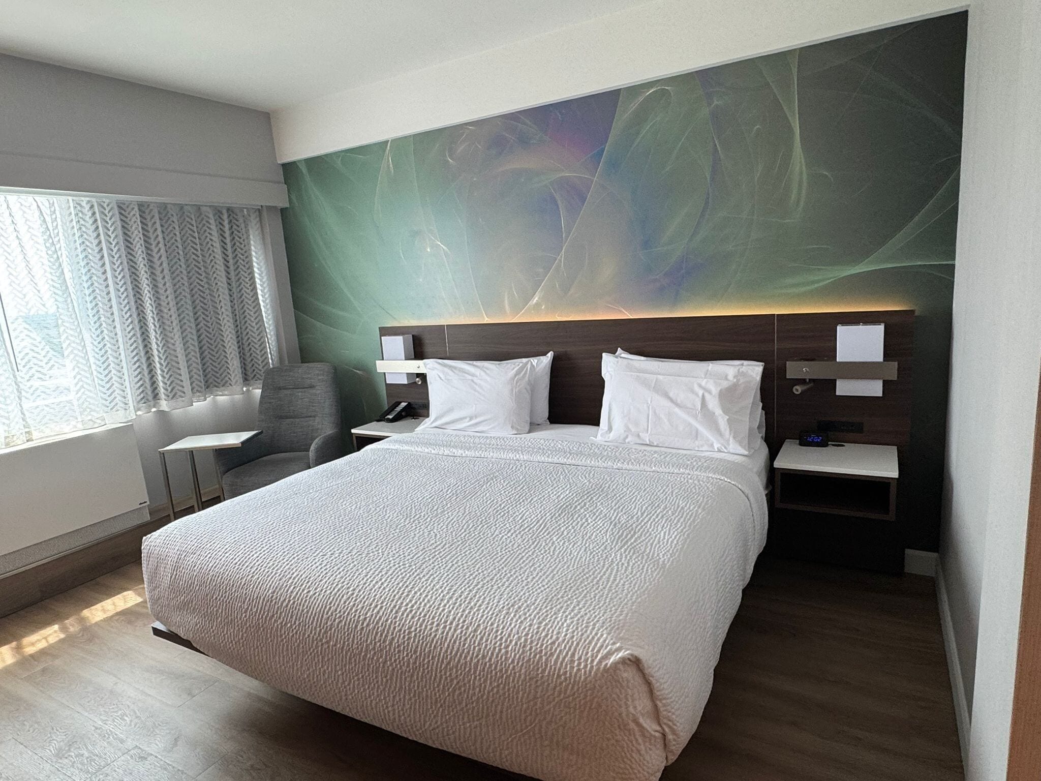A modern hotel room with a large bed, two bedside tables, a chair, and a view of the outdoors through sheer curtains.