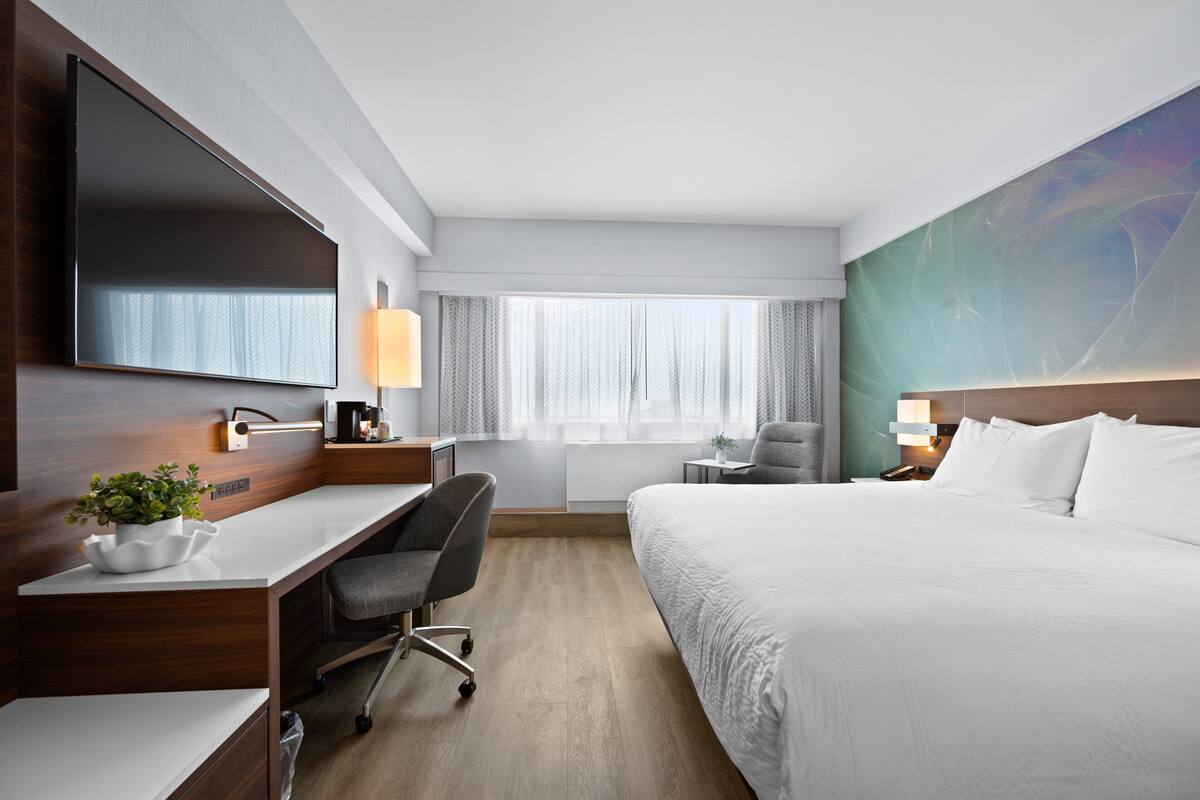 Habitación de hotel con una cama grande, un escritorio con una silla, un televisor de pantalla plana, una lámpara y una ventana con cortinas.