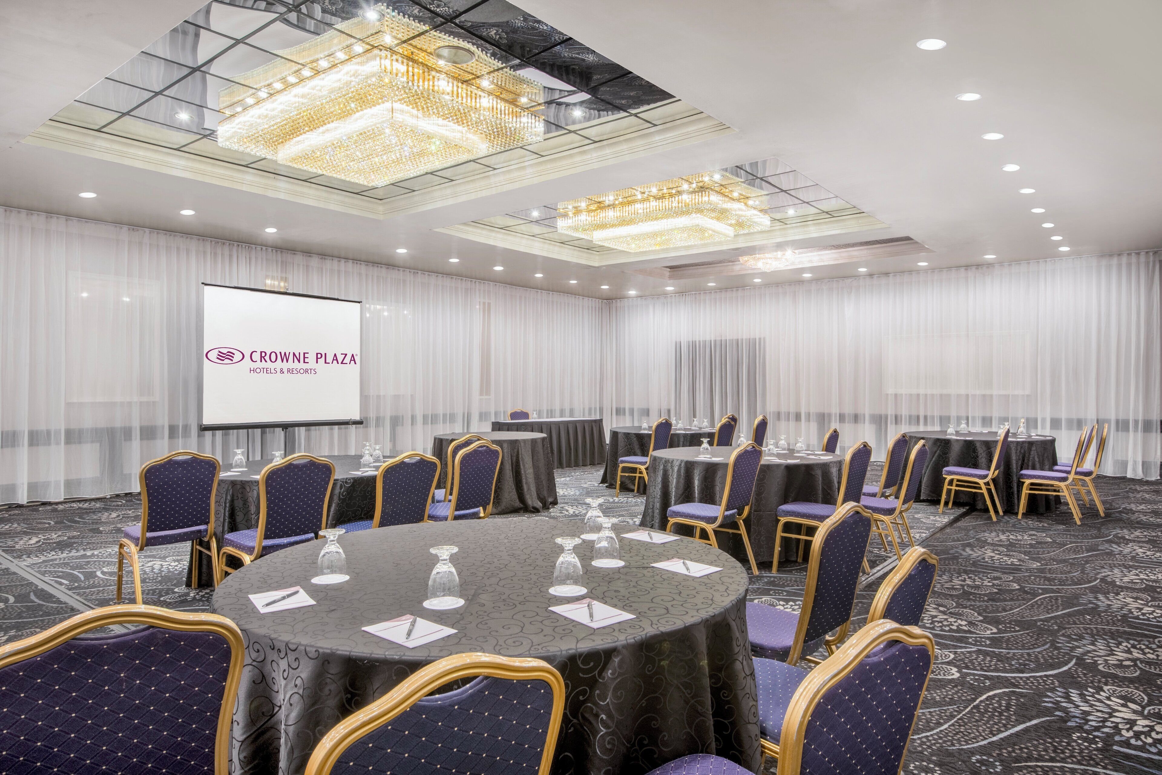 Une salle de conférence équipée de tables rondes, de chaises violettes et d’un grand écran affichant le logo de la chaîne Crowne Plaza Hotels & Resorts.