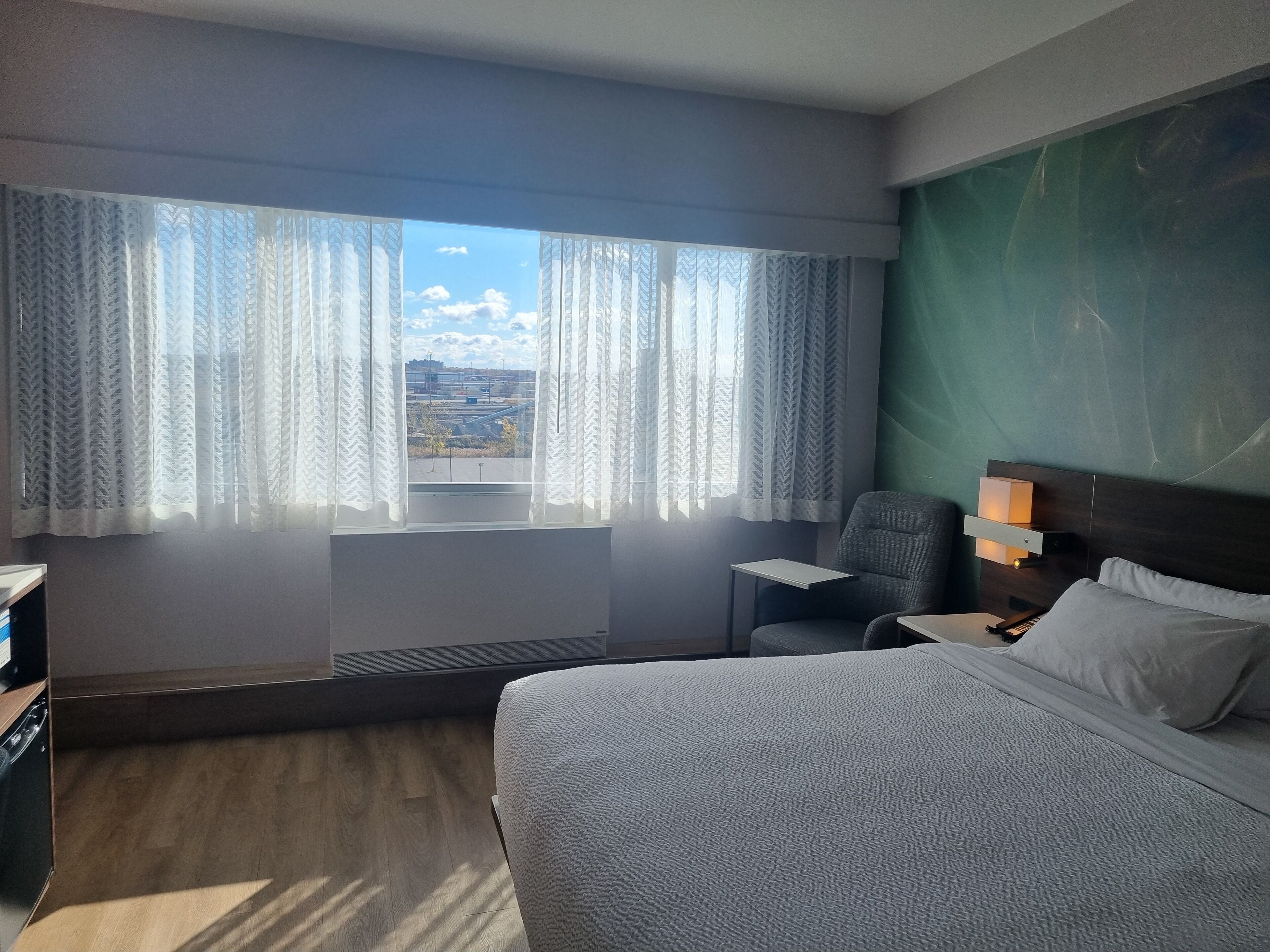 A hotel room with a bed, a chair, a desk, and a view of the cityscape through large windows with sheer curtains.