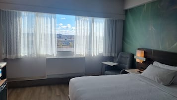 Une chambre d’hôtel avec un lit, une chaise, un bureau et une vue sur le paysage urbain, grâce à de grandes fenêtres ornées de rideaux légers.