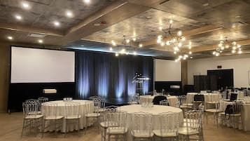 A conference room with round tables, clear chairs, a stage with curtains, and a large screen.