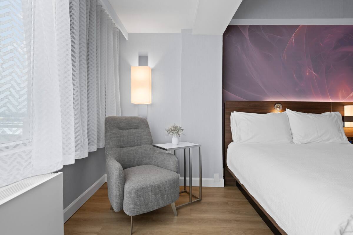 Una habitación de hotel con una cama, un sillón gris, una mesita con una lámpara y un adorno en la pared.
