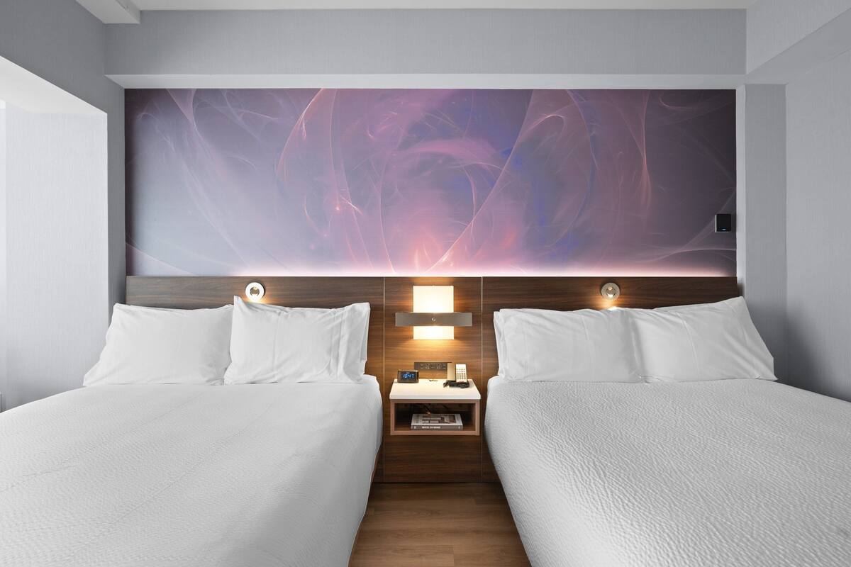 Una habitación de hotel con dos camas, una mesita de noche con lámpara y un panel de control en la pared.
