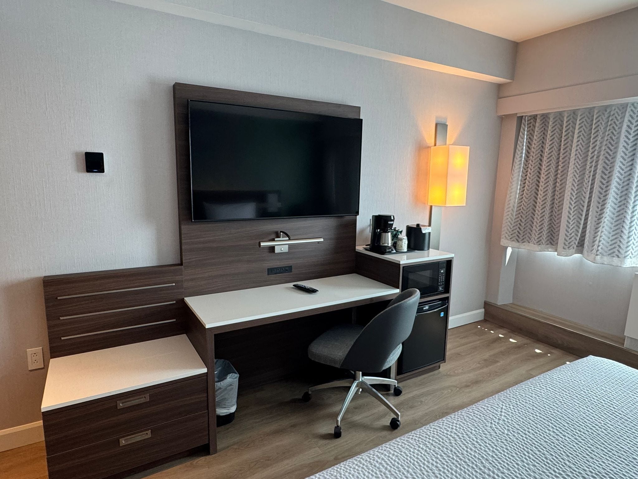 A hotel room with a flat-screen TV, a desk with a chair, a coffee maker, and a lamp.