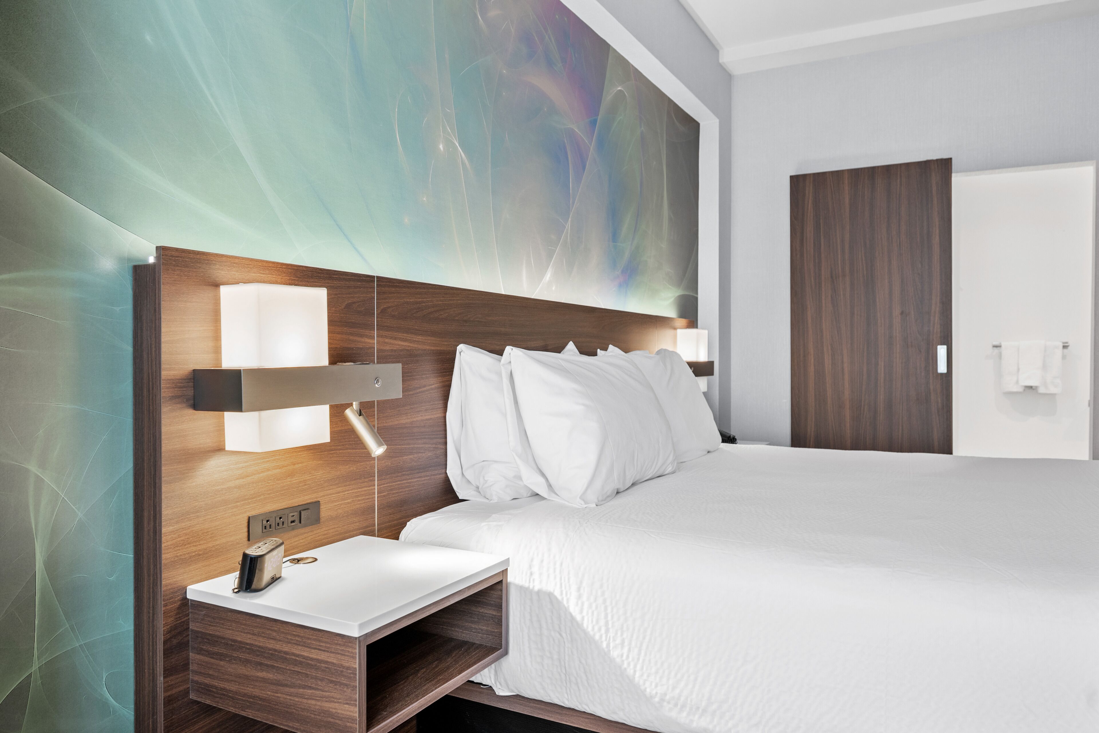 A modern hotel room with a large bed, a bedside table with a lamp, and a wall-mounted light fixture.