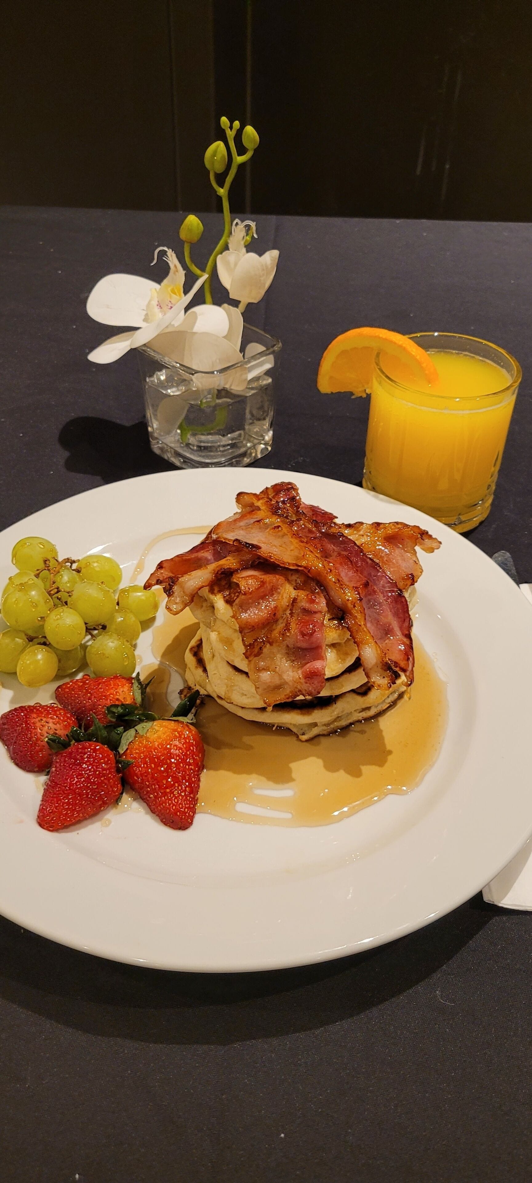 Une assiette de petit-déjeuner comprenant des crêpes, du bacon, des fraises et des raisins, accompagnée d’un verre de jus d’orange et d’un petit vase avec des fleurs blanches.