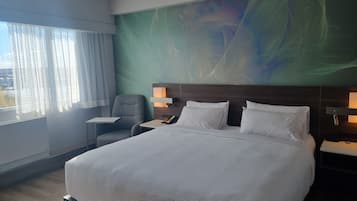 A hotel room with a large bed, a chair, a bedside table, and a nightstand with a lamp.