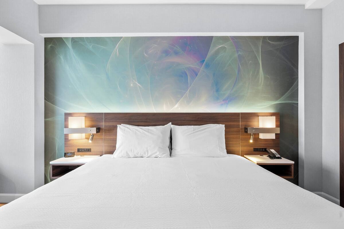 Una habitación de hotel moderna con una cama grande, dos mesitas de noche con lámparas y una obra de arte colgada en la pared sobre la cama.