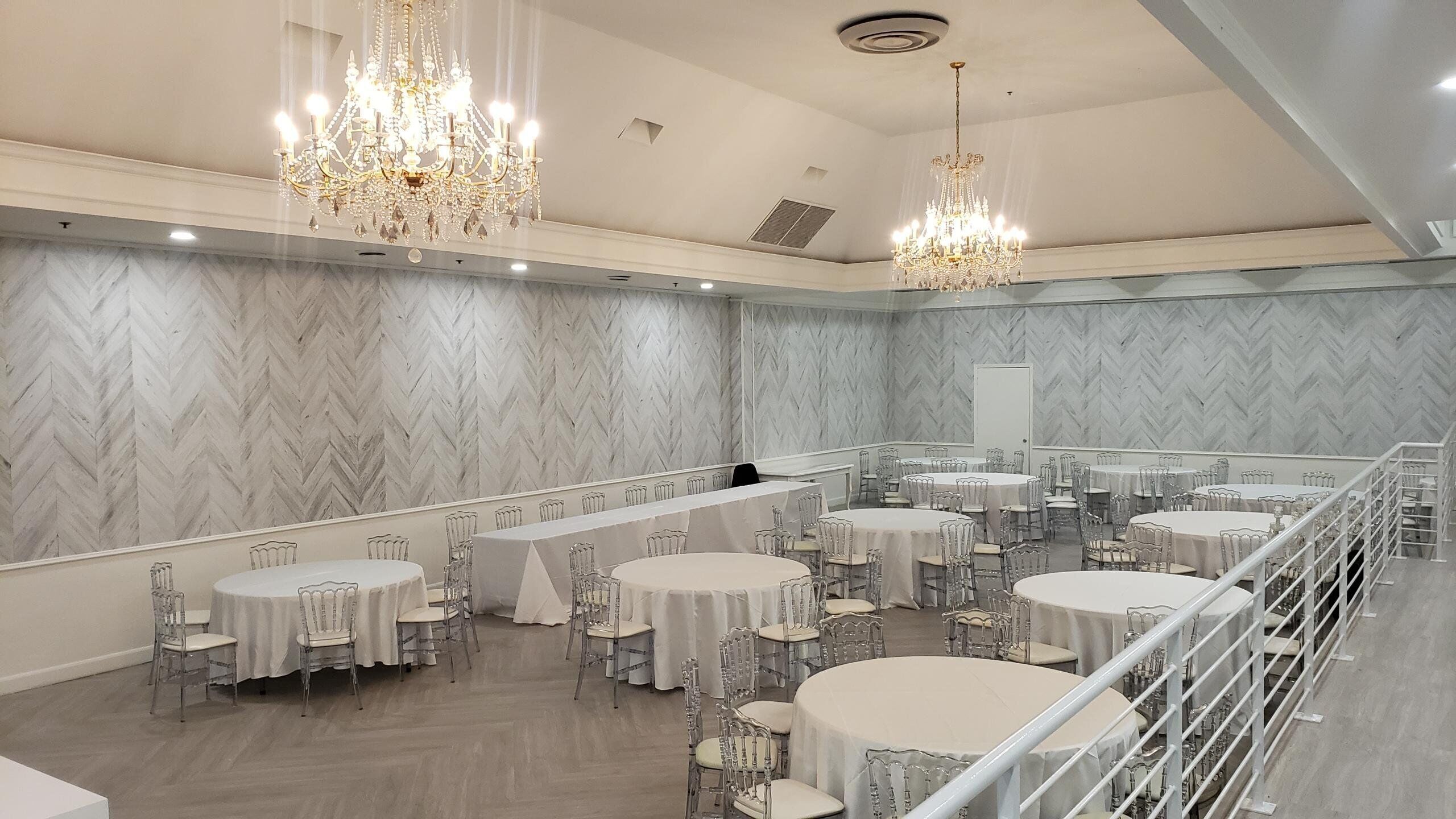 A spacious banquet hall with multiple round tables and chairs, elegant chandeliers, and marble-patterned walls.